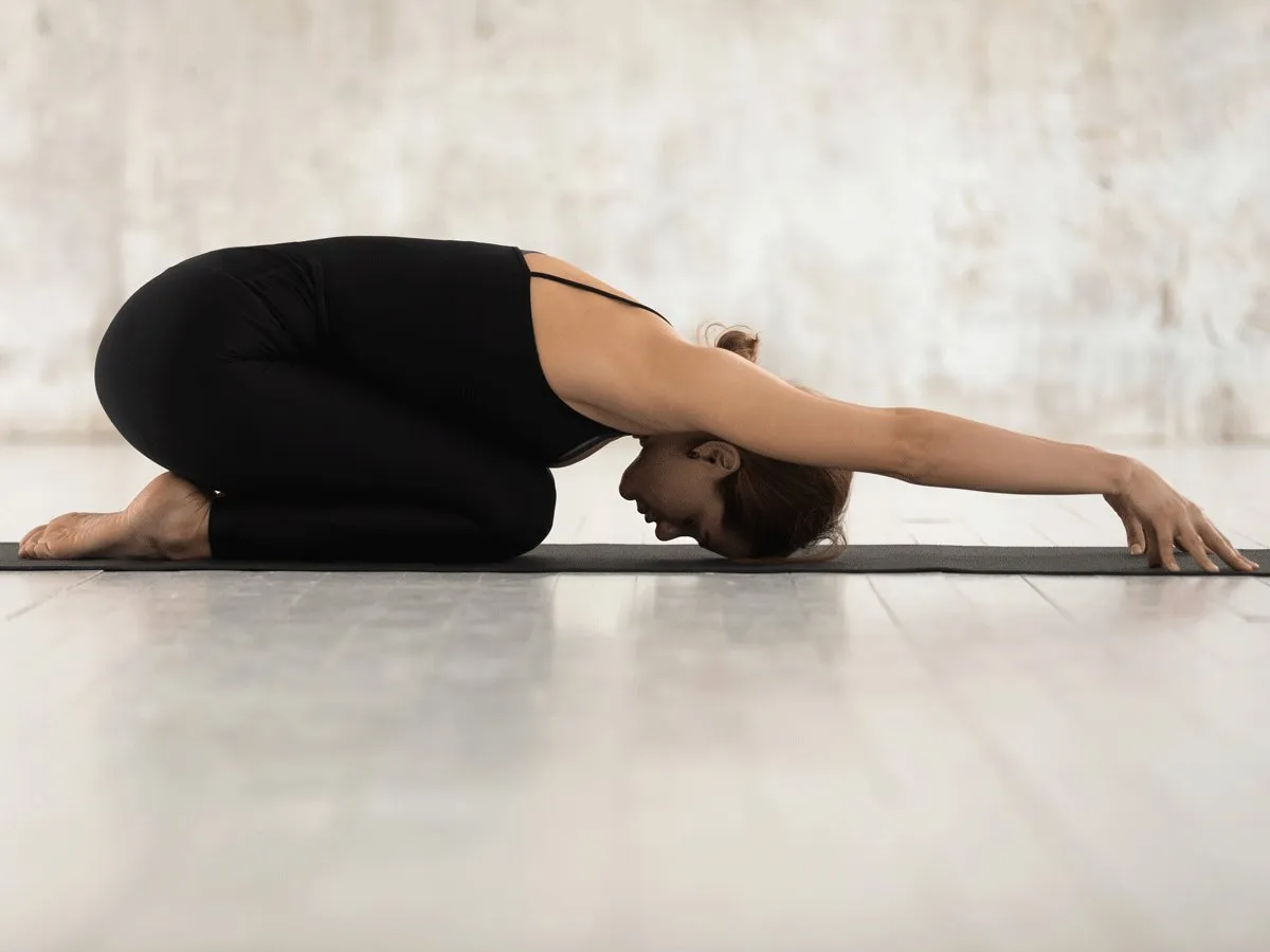 A woman practicing yoga is shown in the Balasana pose (Child’s Pose), resting on a black yoga mat placed on a light-colored wooden floor. She is kneeling with her legs folded under her, hips sitting back on her heels, and torso gently resting over her thighs. Her forehead touches the mat while her arms are extended straight forward with palms facing down, creating a sense of deep relaxation and surrender. She is dressed in a black, fitted yoga outfit for flexibility that allows for unrestricted movement. The background is minimalistic, with a soft, blurred neutral wall that keeps the focus on the pose. The lighting is natural and soft, contributing to the serene and meditative atmosphere of the scene—ideal for those seeking gentle yoga for menstrual cramps, yoga for period pain relief, or restorative yoga poses for women’s health. This image of Balasana (Child’s Pose) reflects a natural method for relieving period pain which supports the campaign "STOP The Period Pain" which is a knowledge initiative campaign by Blue Cross Laboratories, the makers of Meftal Spas.